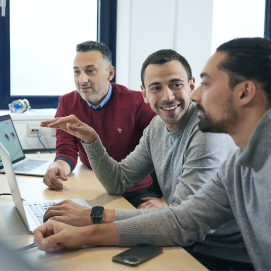 Three men are seated closely together in front of a laptop, engaged in a lively discussion about the content displayed on the screen. Their expressions range from smiles to looks of keen interest, suggesting a positive and collaborative atmosphere. The shared focus on the laptop screen indicates they are likely working on a project or solving a problem together.