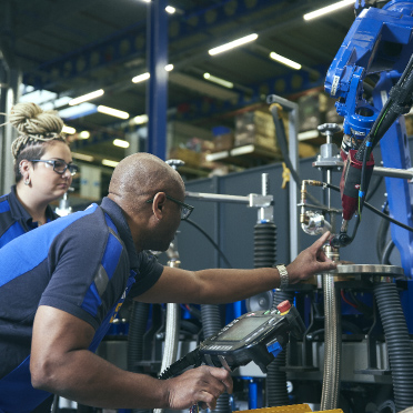 An image depicts a man and a woman collaboratively working on an advanced machine within the bustling environment of a warehouse. Both individuals are actively engaged, perhaps adjusting settings, analyzing data on a monitor, or performing maintenance, showcasing their teamwork and technical proficiency.