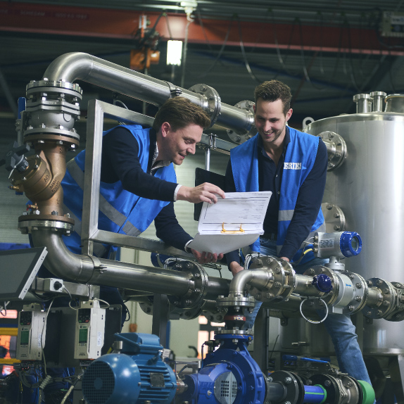 Two young men, both wearing blue ERIKS vests, share a moment of camaraderie and collaboration in a warehouse setting. They are smiling and talking, deeply engaged in working on a project together. The background is adorned with an assortment of metal pipes and various types of hoses, adding a layer of industrial ambiance to the scene. This image vividly captures the spirit of teamwork and the hands-on approach to work within the context of an organized, well-stocked warehouse environment.